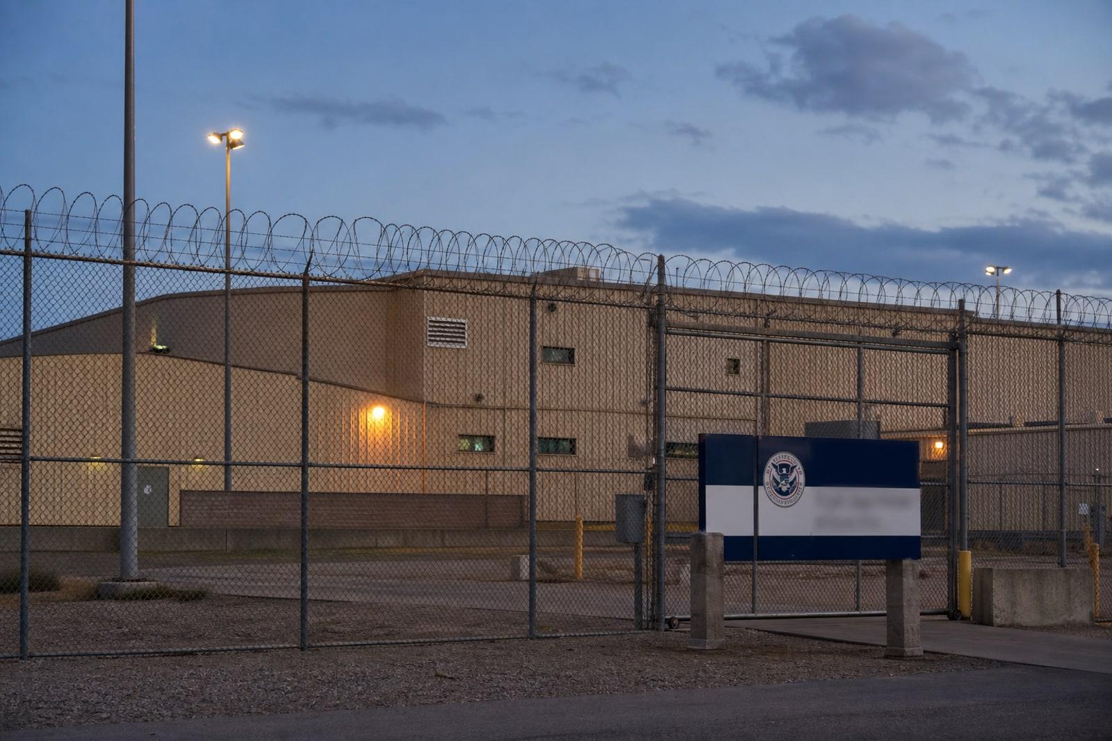 The razor-wire-topped chain-link perimeter fence of a US ICE immigration detention facility with a low institutional building behind it.