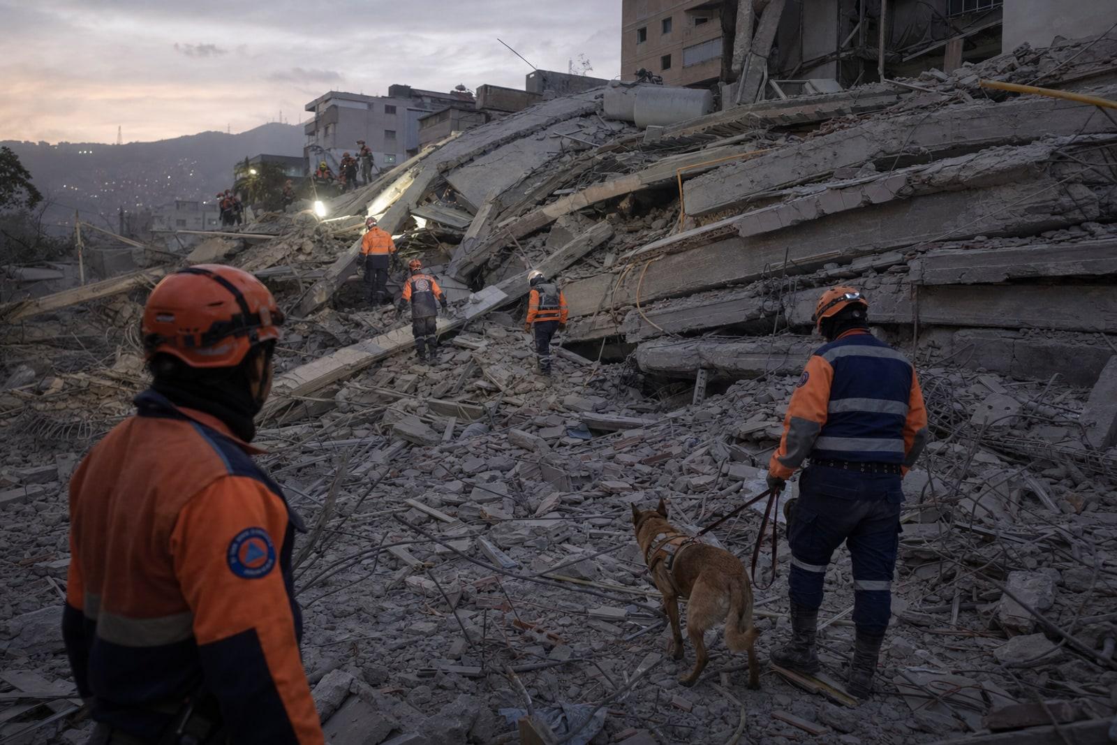 Search-and-rescue workers in high-visibility gear and a dog searching the rubble of a collapsed concrete apartment building in Caracas at dusk.