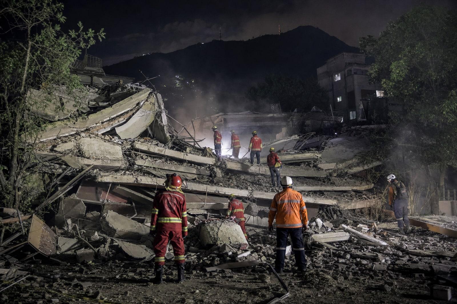 Rescue workers in orange and red gear search the rubble of a collapsed mid-rise apartment building in Caracas at night under floodlights.