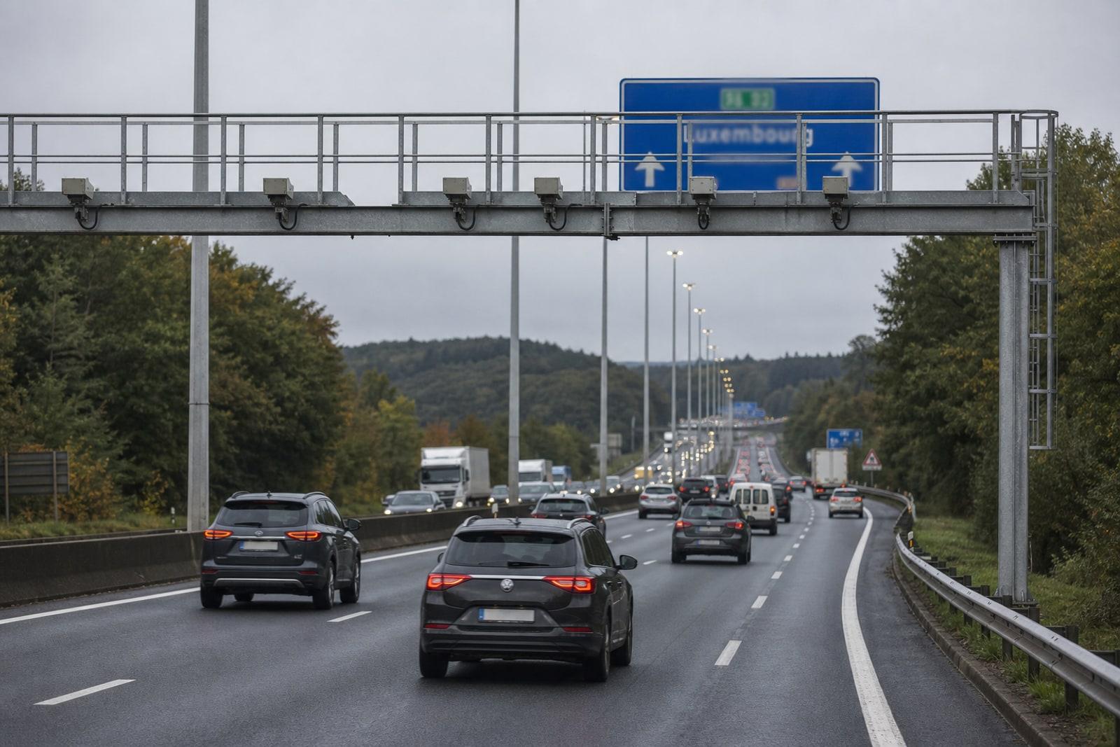 Autoen am Pendlerverkéier op der E411 an der Belsch, ënner engem Portal mat Kameraen an engem bloen Autobunnsschëld Richtung Lëtzebuerg.