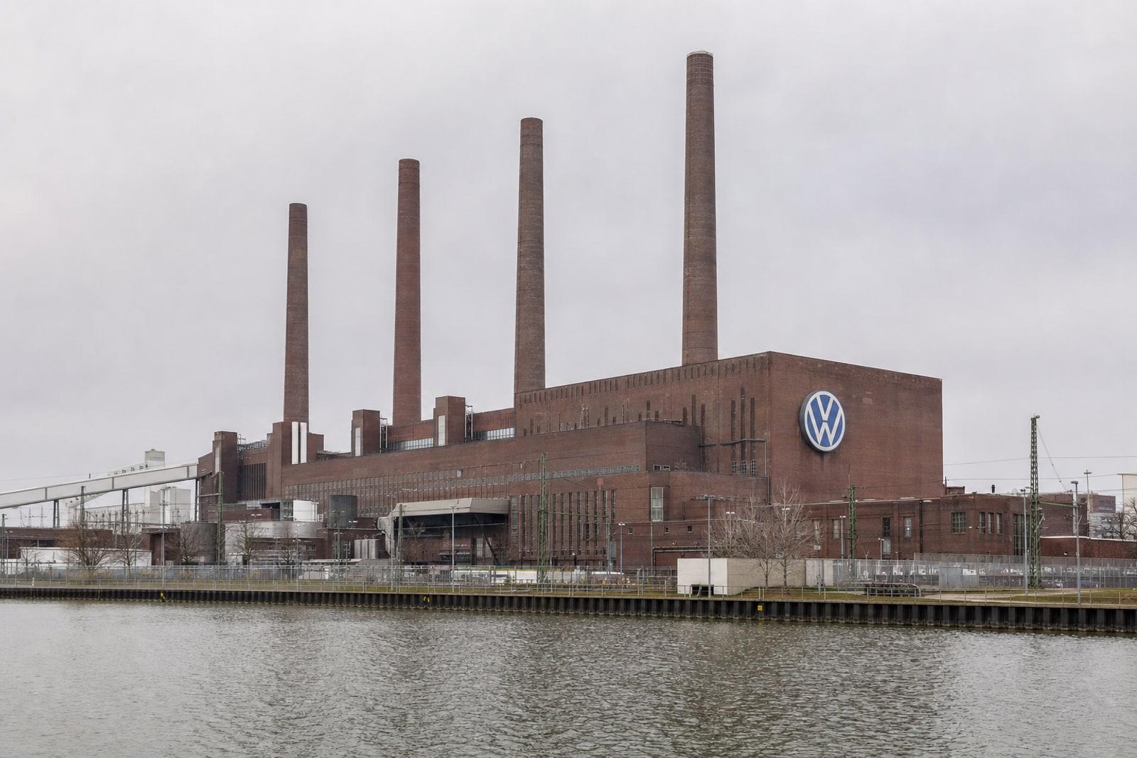 Volkswagen's Wolfsburg plant with its four red-brick power-station chimneys and the blue-and-white VW roundel under an overcast sky.